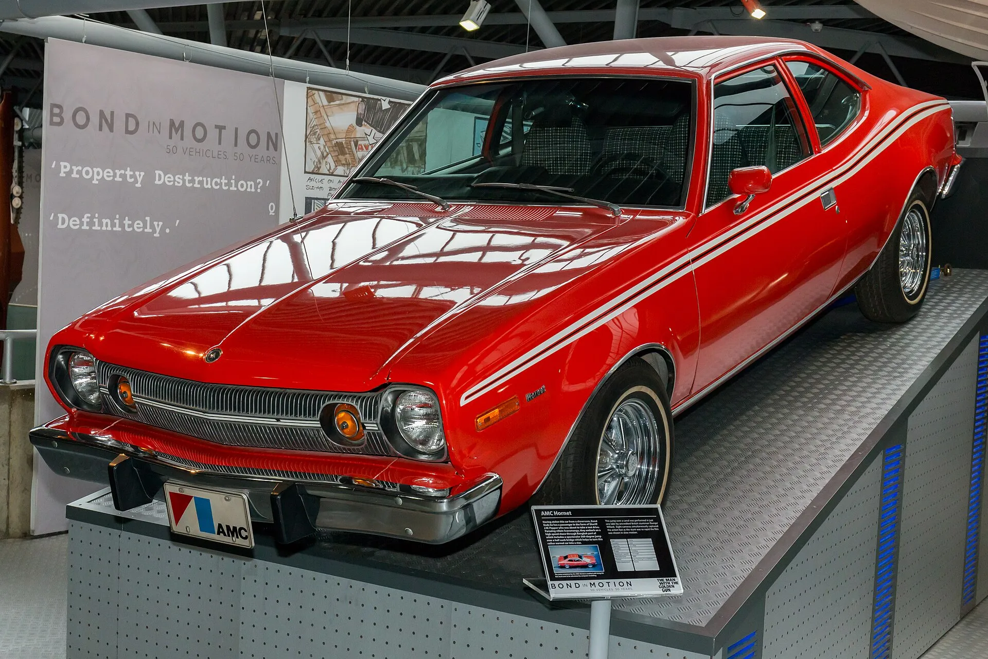 Roter AMC Hornet Hatchback mit weißen Doppelstreifen und verchromten Speichenrädern auf einem Podest, dahinter ein Banner der Ausstellung „Bond in Motion“