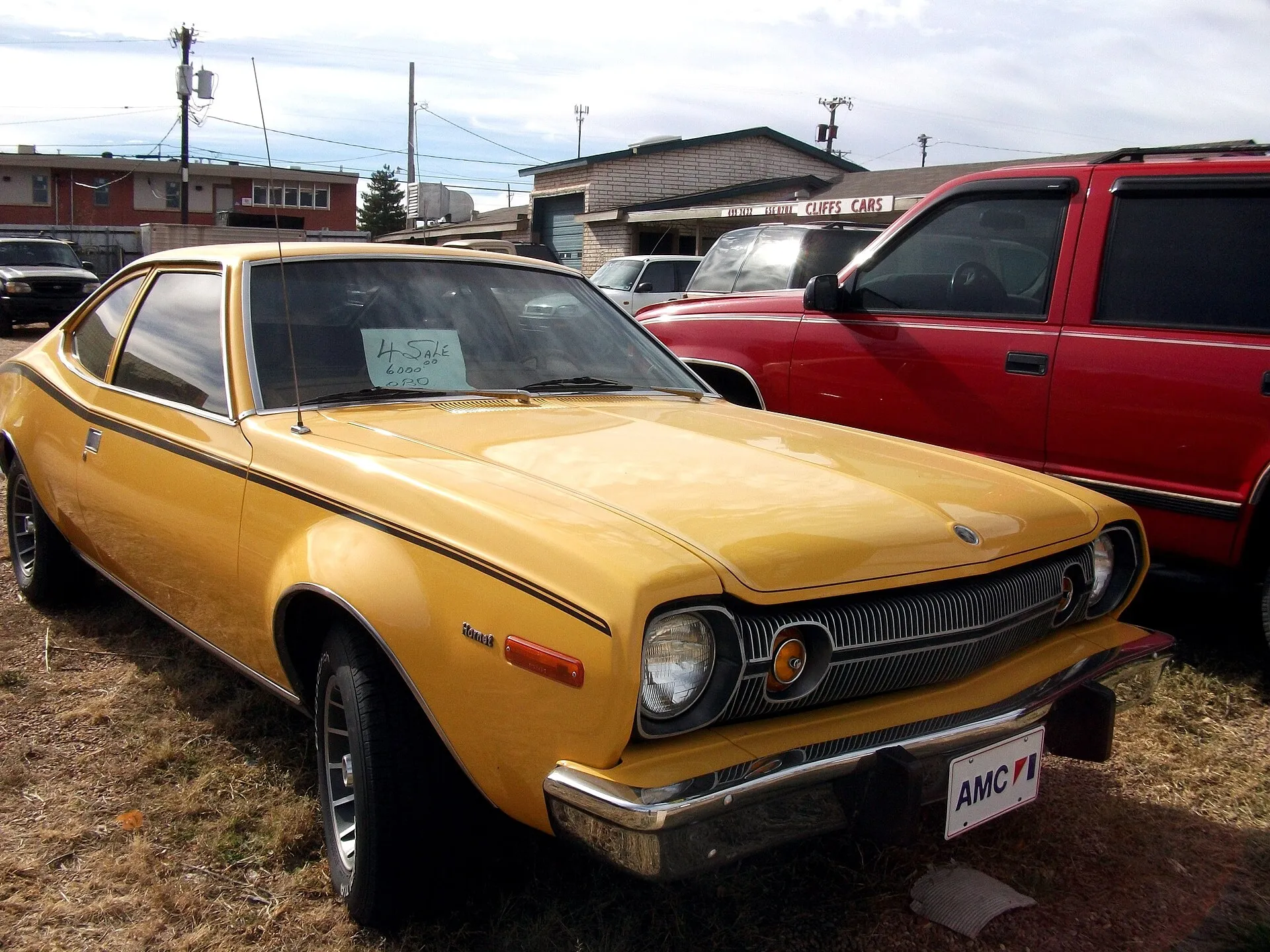 Ein ockergelber AMC Hornet Hatchback des Baujahrs 1974 in Dreiviertelansicht von vorn auf einem Gebrauchtwagenplatz