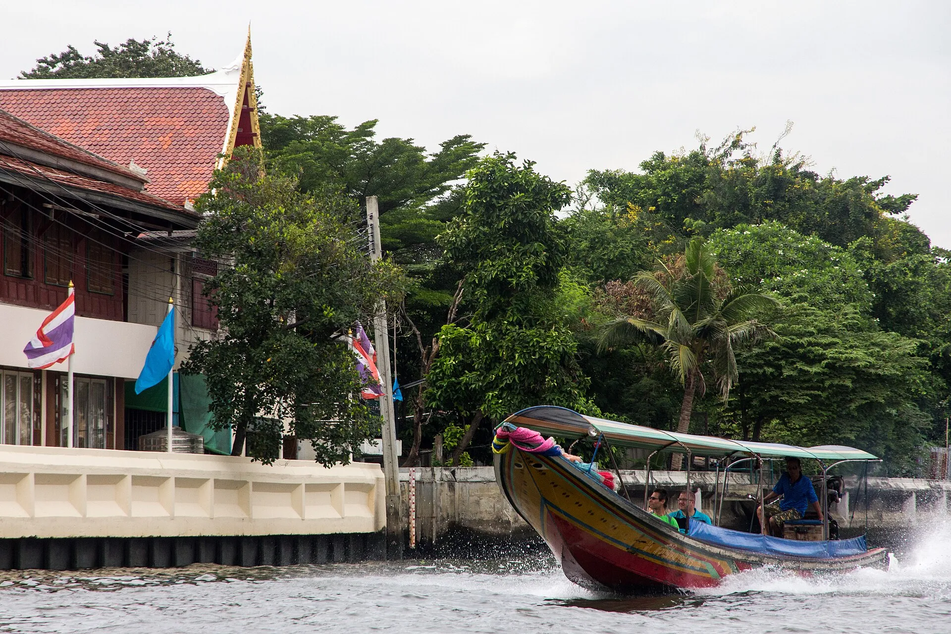 Ein besetztes Longtailboot fährt einen Kanal in Bangkok hinunter, am Ufer ein Holzgebäude mit rotem Ziegeldach, thailändische Flaggen und dichtes Grün