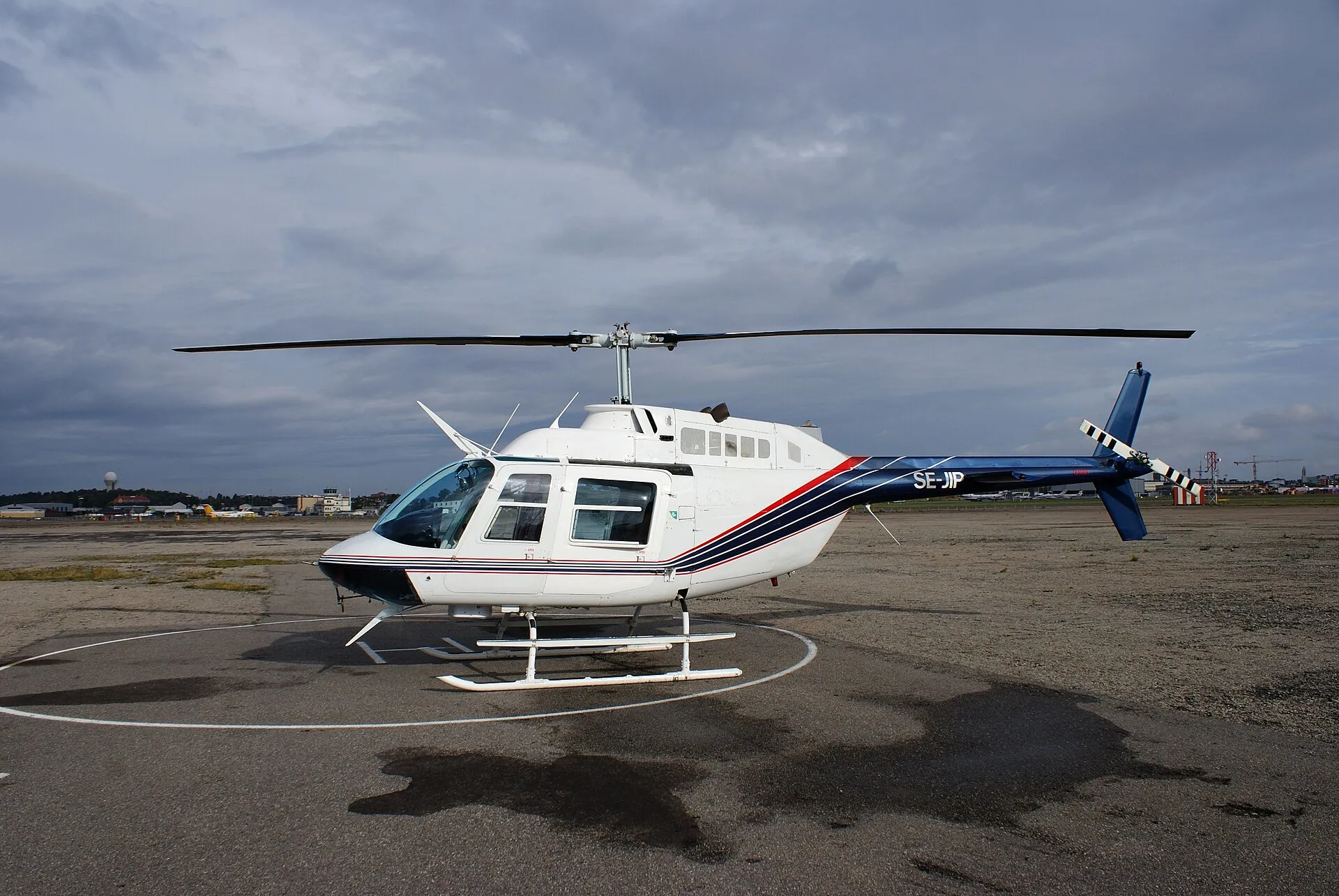Weißer Bell 206 JetRanger mit blau-rotem Zierstreifen auf einem Landeplatz unter bewölktem Himmel