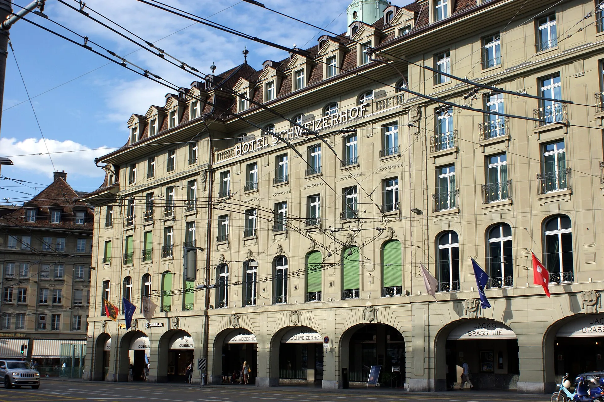 Die Sandsteinfassade des Hotels Schweizerhof in Bern mit Arkaden, Flaggen und Tramoberleitungen davor