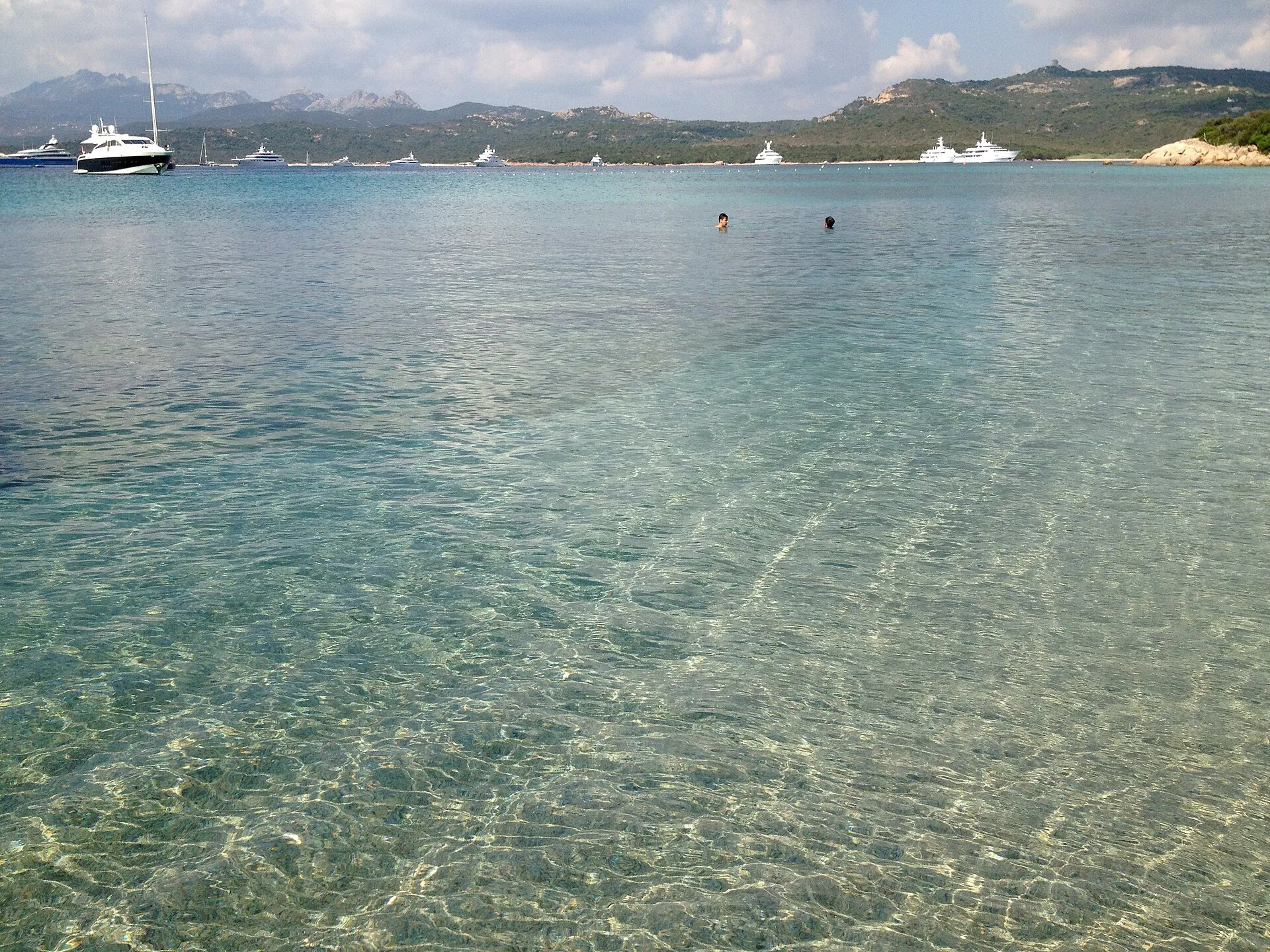 Flaches, klares Wasser am Strand von Capriccioli auf Sardinien mit Jachten und Hügeln im Hintergrund