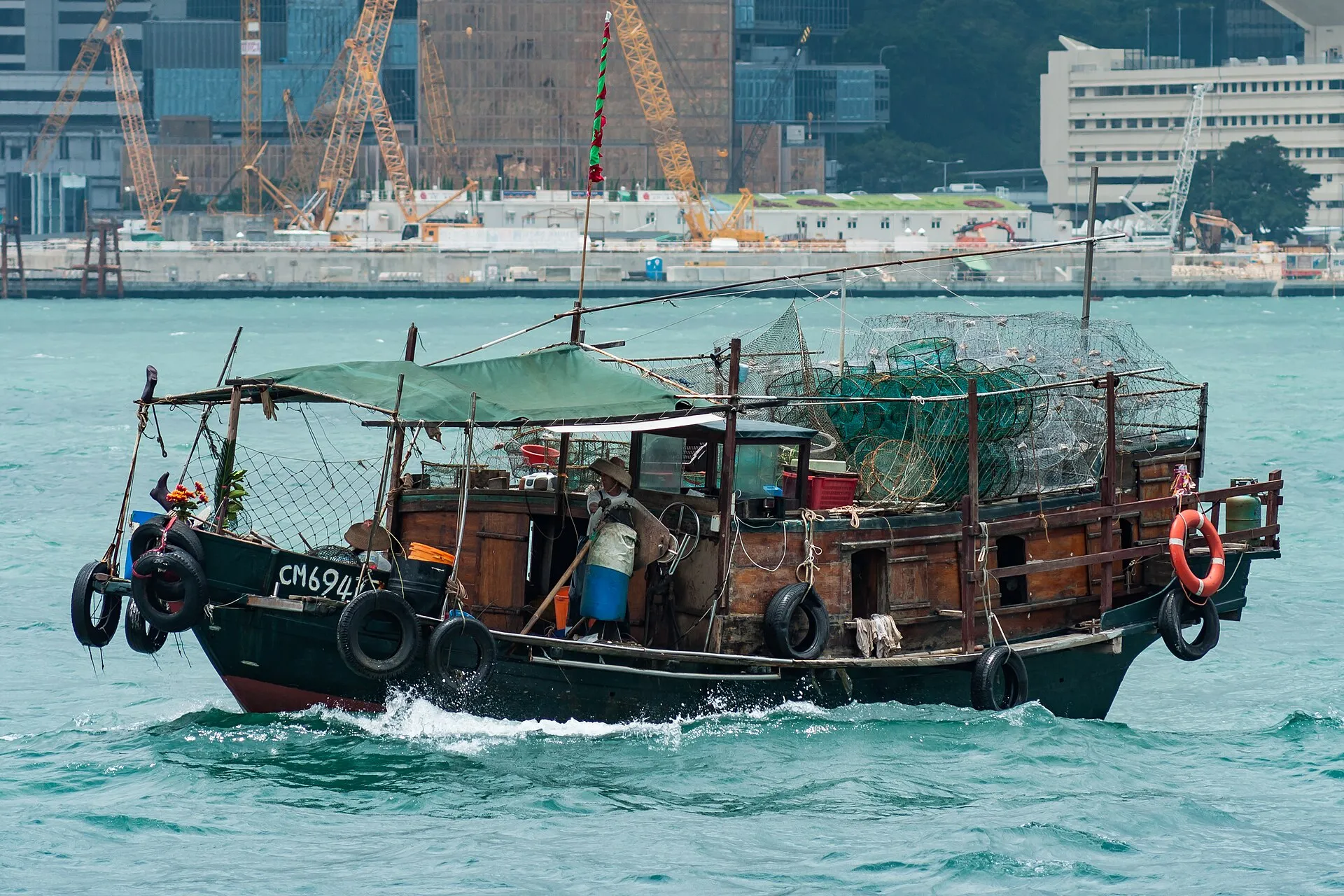 Eine hölzerne chinesische Fischerdschunke mit gestapelten Reusen an Deck fährt durch den Victoria Harbour, dahinter Kräne und Gebäude von Hongkong