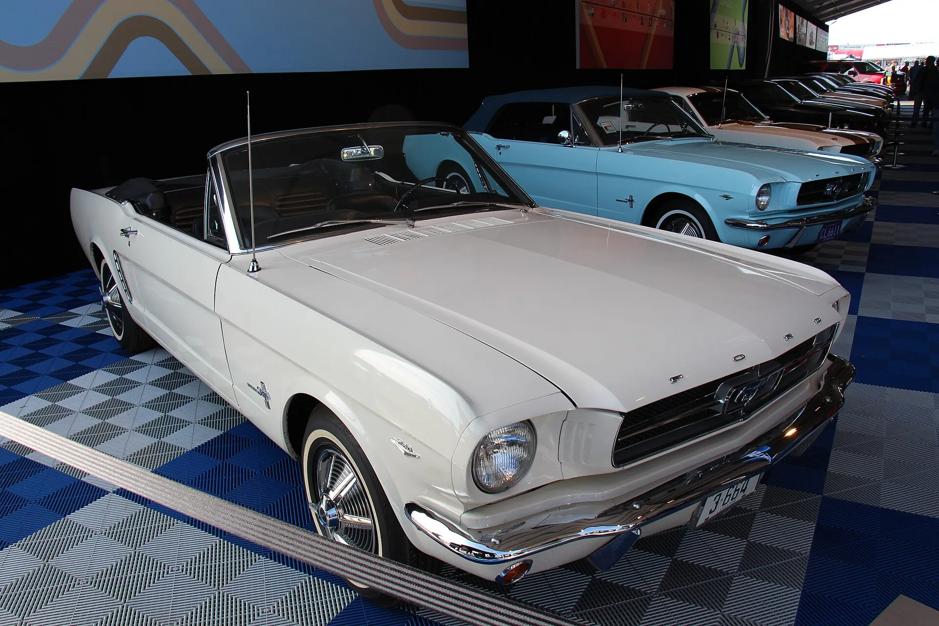 Cremeweißes Ford Mustang Cabriolet von 1964 mit rotem Interieur in einer Ausstellungshalle, dahinter ein hellblauer Mustang