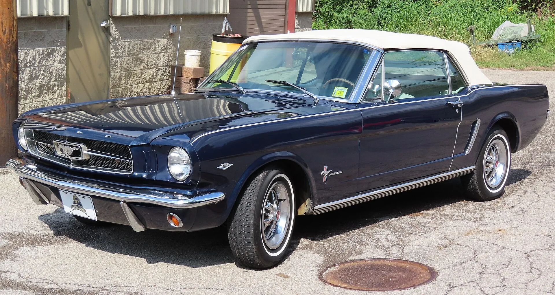 Dunkelblaues Ford Mustang Cabriolet von 1965 mit weißem Verdeck vor einem Werkstattgebäude
