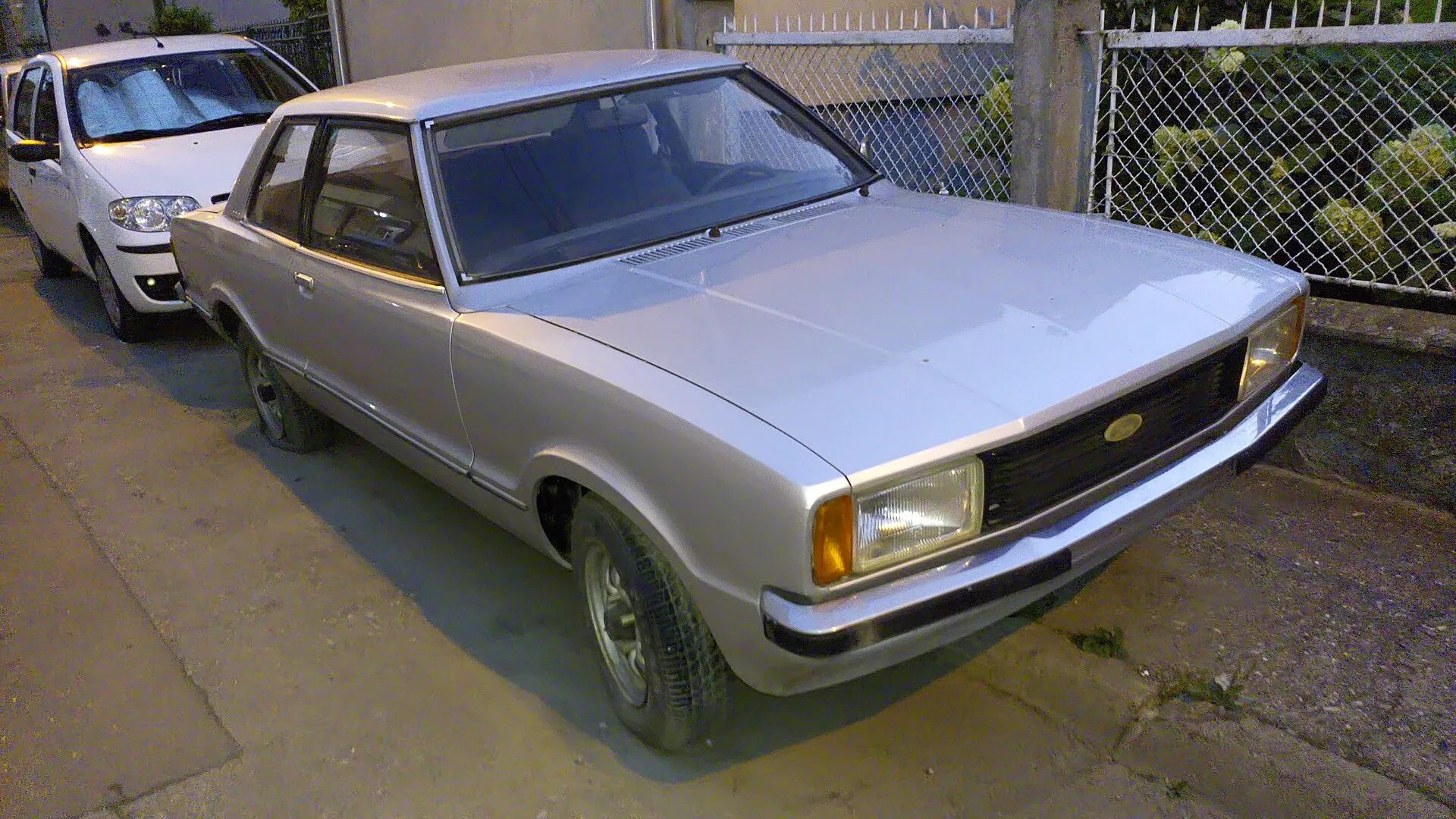 Silberner zweitüriger Ford Taunus der Baureihe TC2 in der Dämmerung von schräg vorn an einem Zaun