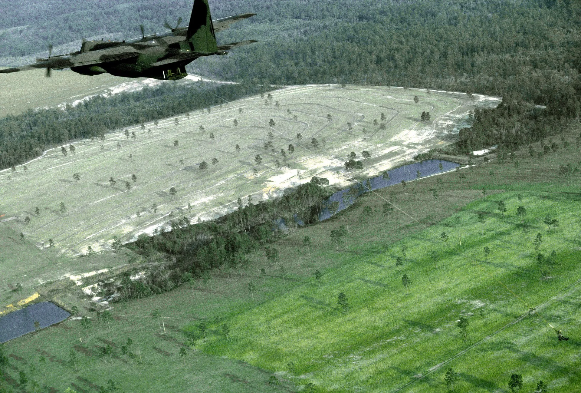 Eine MC-130 Hercules der US Air Force mit ausgeklappter Buggabel überfliegt eine Lichtung; am Boden hängt eine Person an der Leine des Fulton-Systems