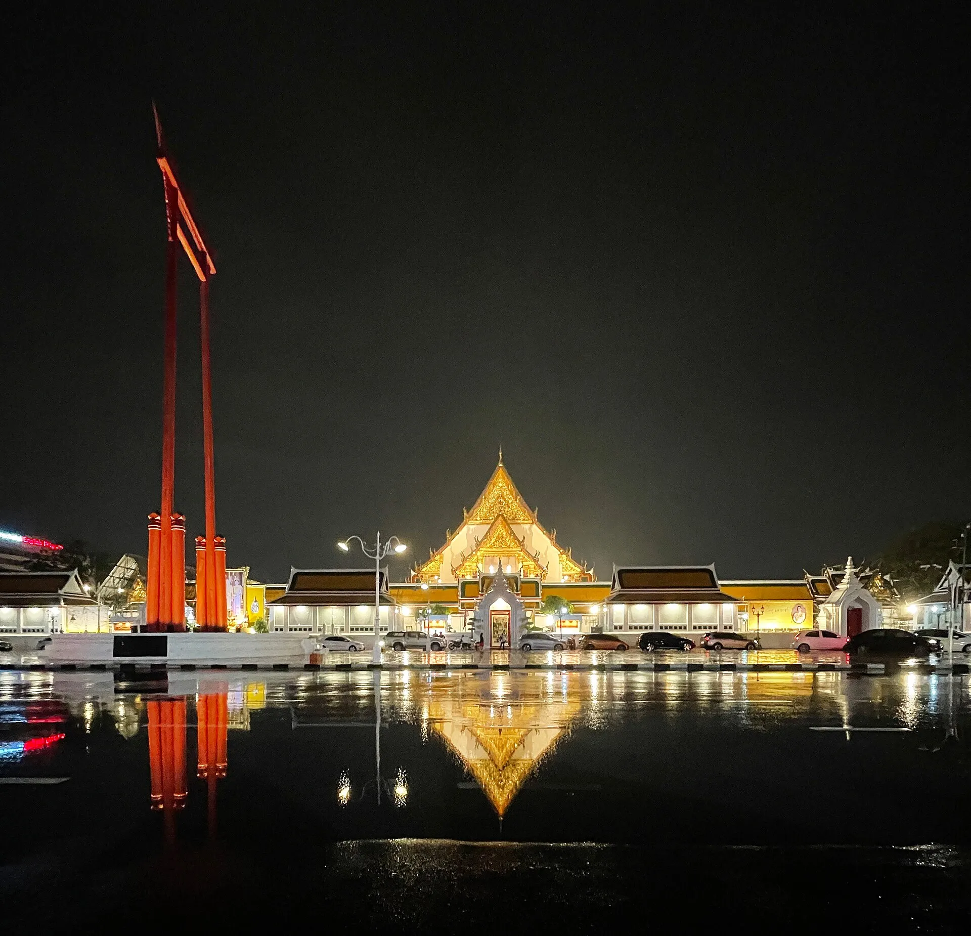 Die rot gestrichene Riesenschaukel Sao Ching Cha und der angestrahlte Tempel Wat Suthat bei Nacht, gespiegelt in der nassen Straße