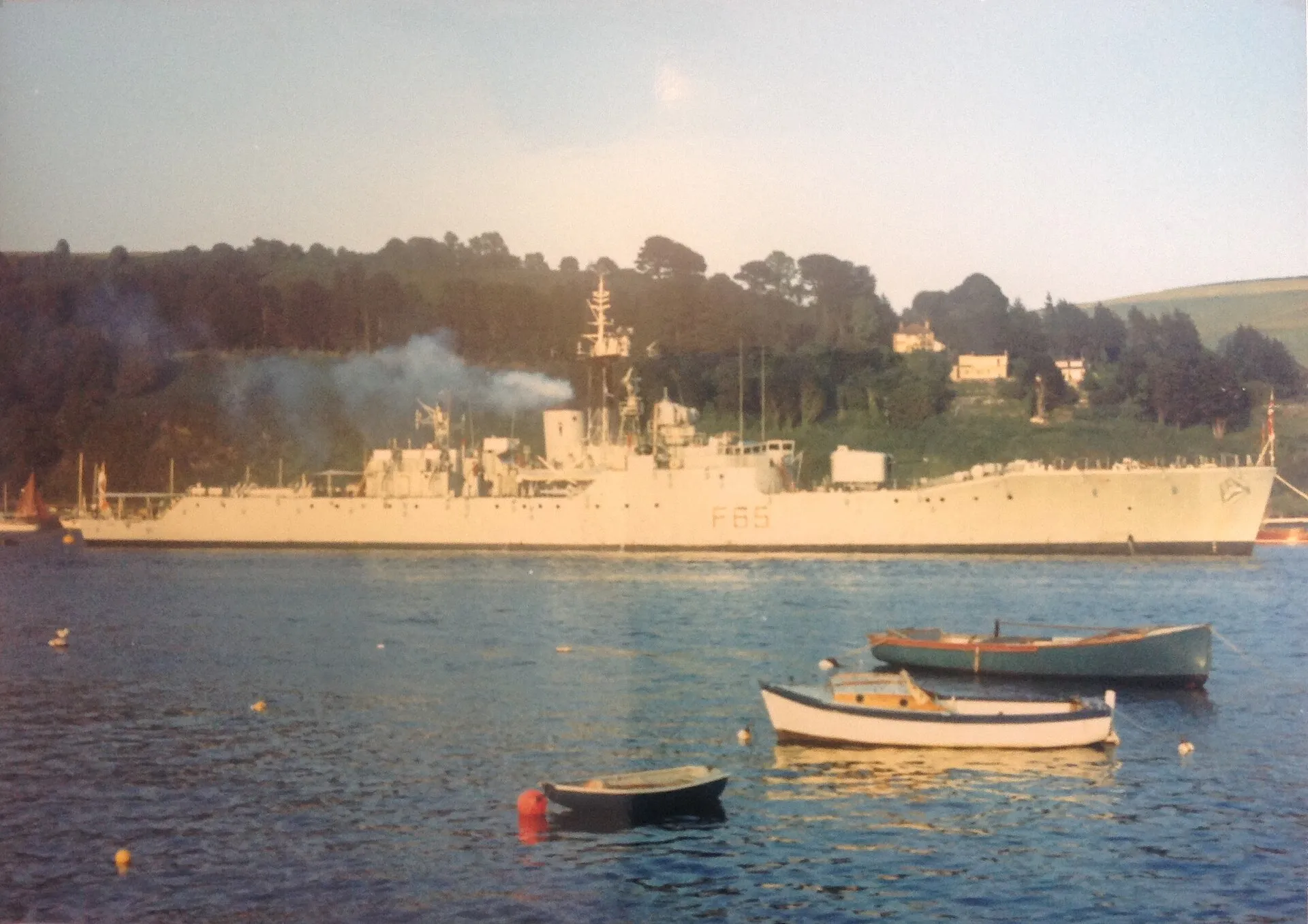 Die graue Fregatte HMS Tenby mit der Kennung F65 vor Anker vor bewaldeten Hügeln, im Vordergrund kleine Boote