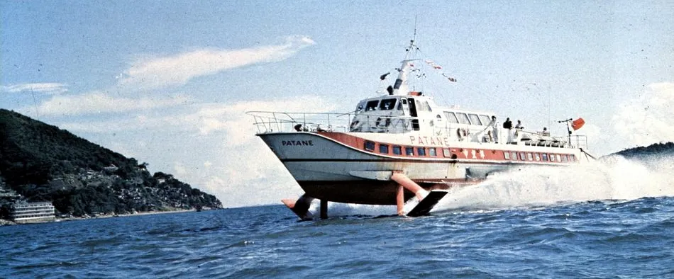Das weiß-rote Tragflügelboot Patane fährt aufgeständert auf seinen Tragflächen über das Meer, im Hintergrund eine bewaldete Küste