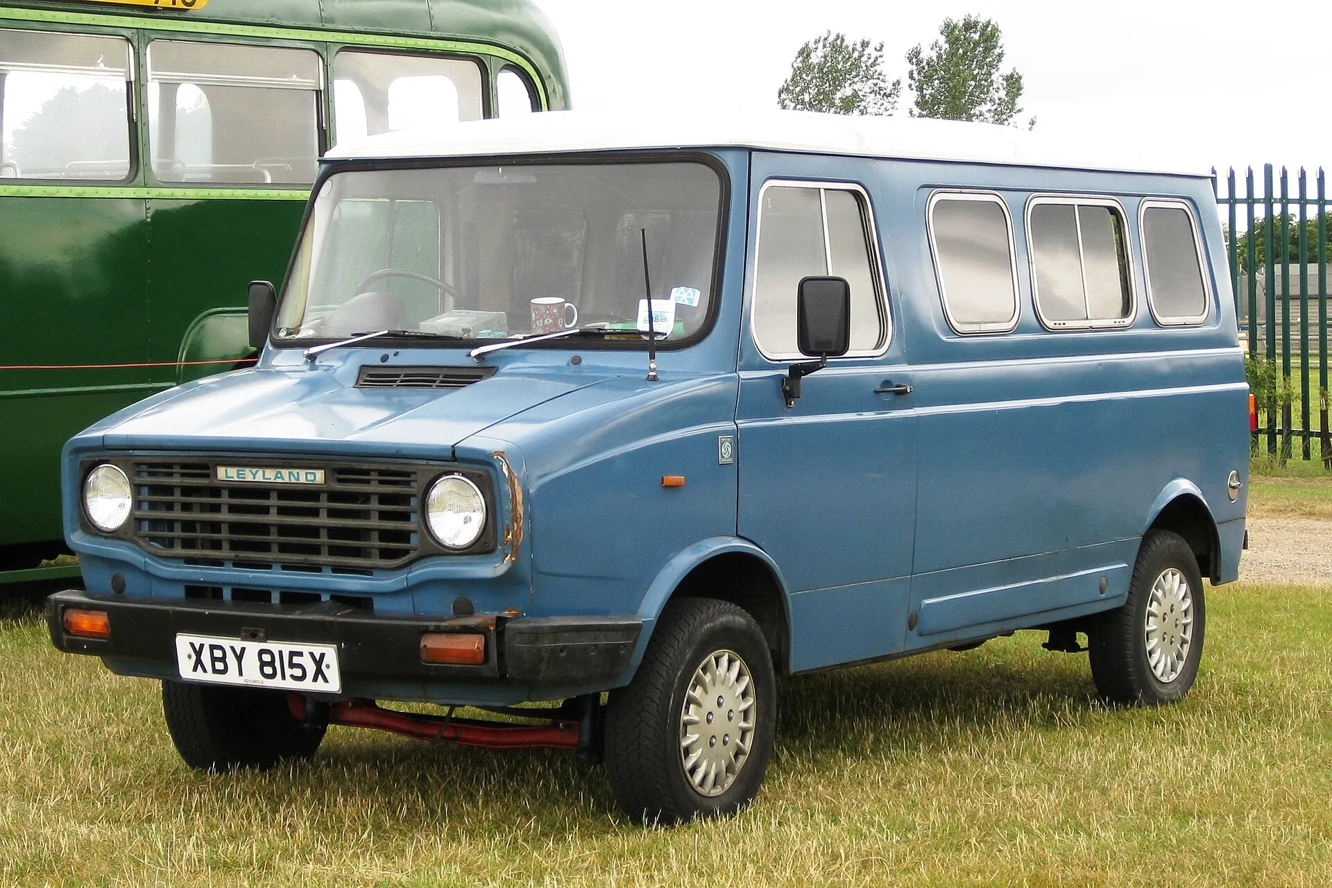 Blauer Leyland Sherpa mit weißem Dach und seitlichen Fenstern auf einer Wiese, dahinter ein grüner Omnibus