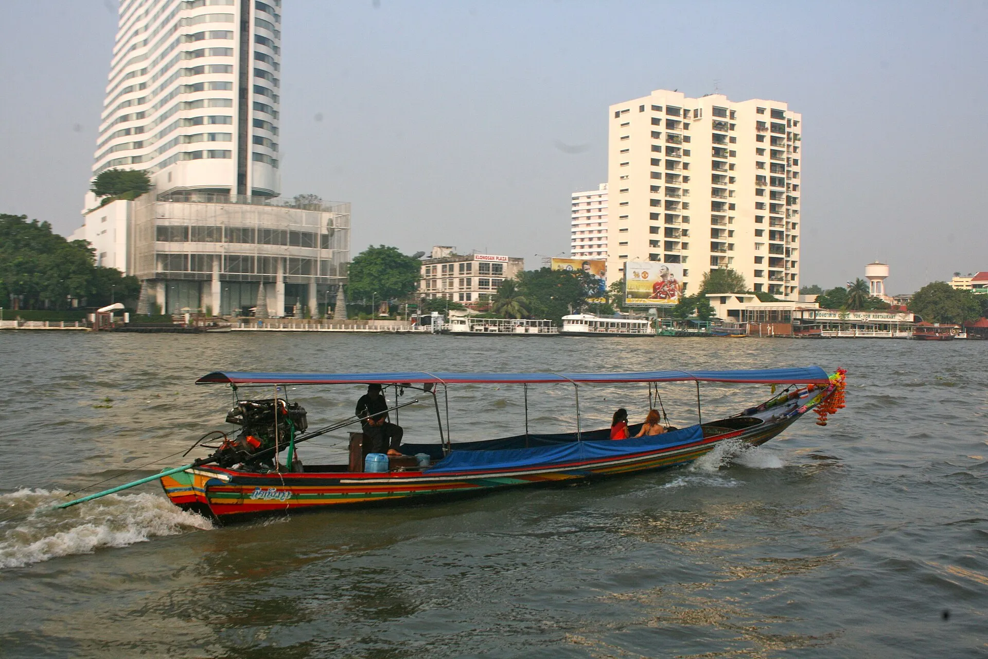 Buntes Longtailboot mit blauem Sonnendach auf dem Chao Phraya in Bangkok; im Heck steht der offene Motor auf einem Drehlager, die lange Welle ragt weit ins Wasser