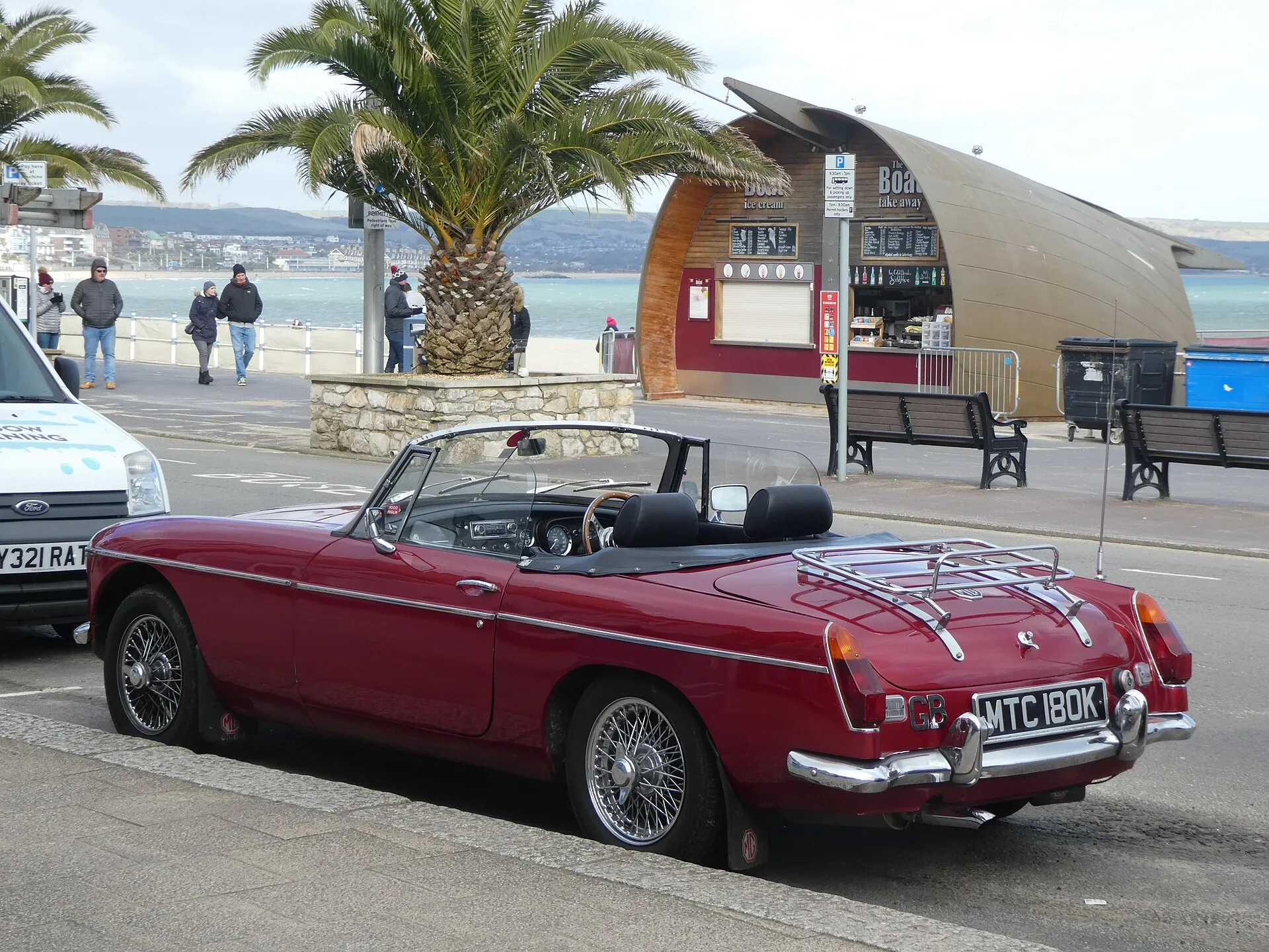 Dunkelroter MGB Roadster von 1972 mit Chromstoßstangen, Speichenrädern und Gepäckträger, schräg von hinten an einer Strandpromenade