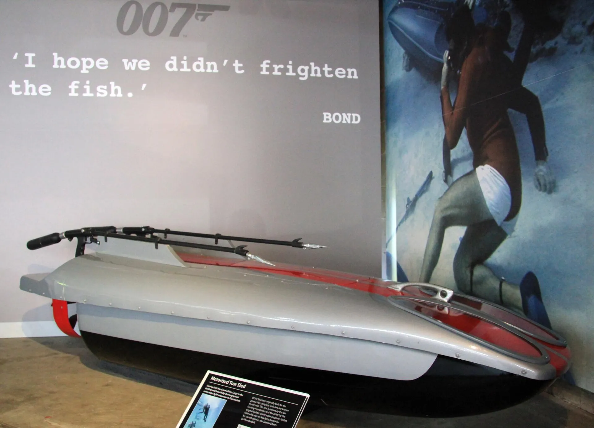 Silber-roter Unterwasserschlitten mit zwei Harpunen in der Ausstellung Bond in Motion, dahinter ein Filmzitat an der Wand