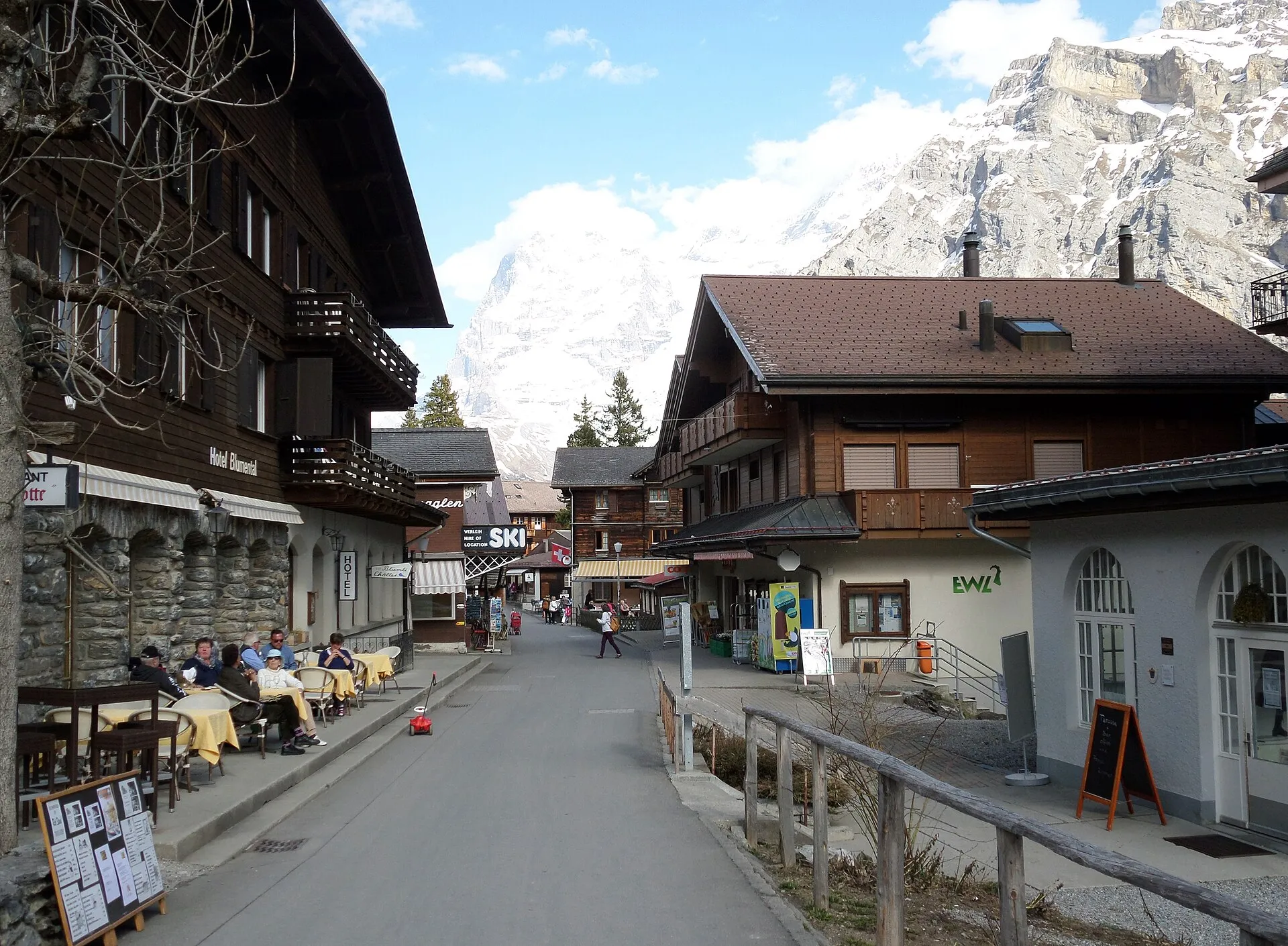 Dorfstraße von Mürren mit Chalets, Hotelterrasse und Skiverleih vor einer steilen Felswand
