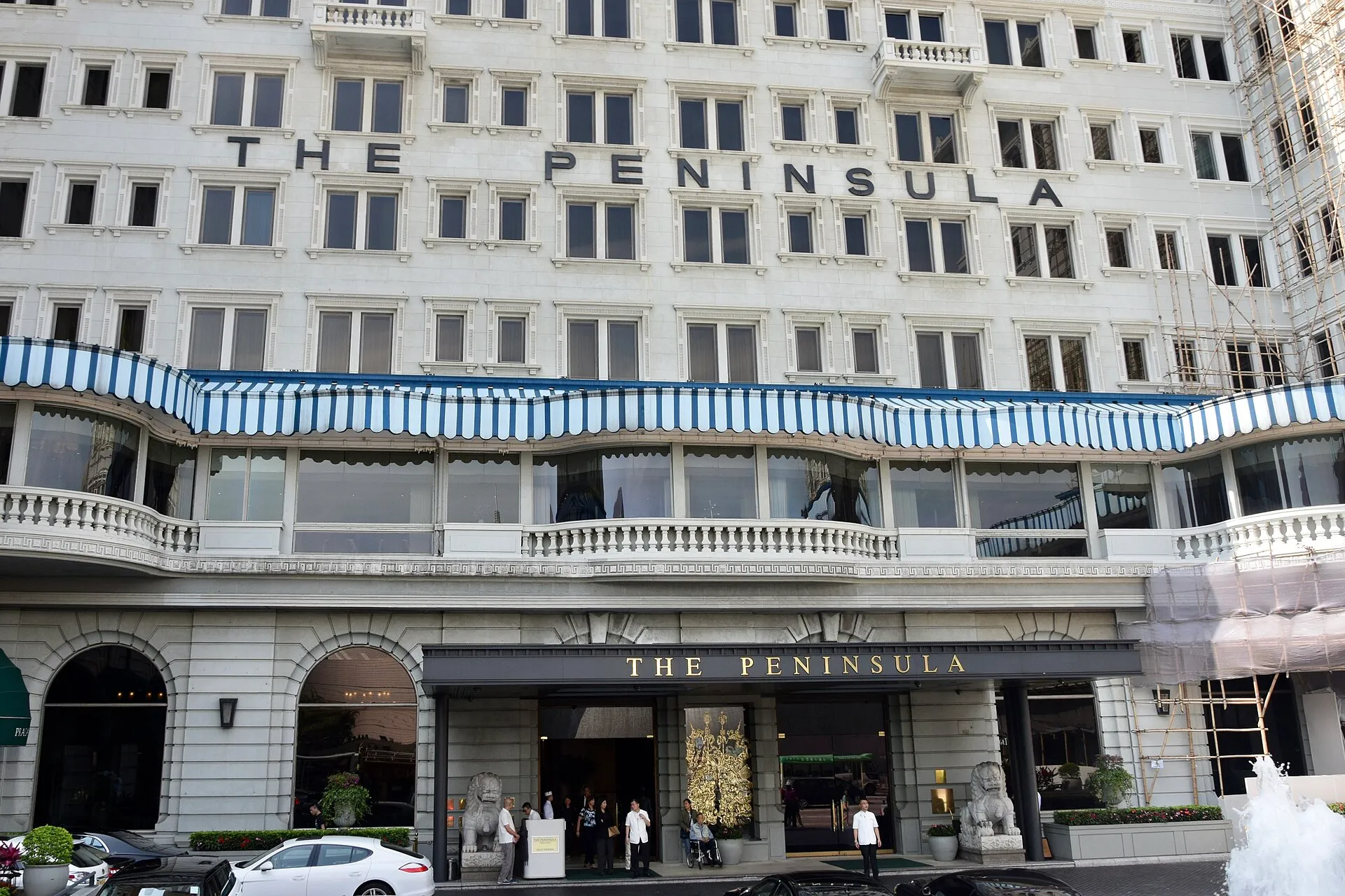 Die weiße Fassade des Hotels The Peninsula in Kowloon mit dem Schriftzug über den blau-weiß gestreiften Markisen, davor die Vorfahrt mit steinernen Löwen