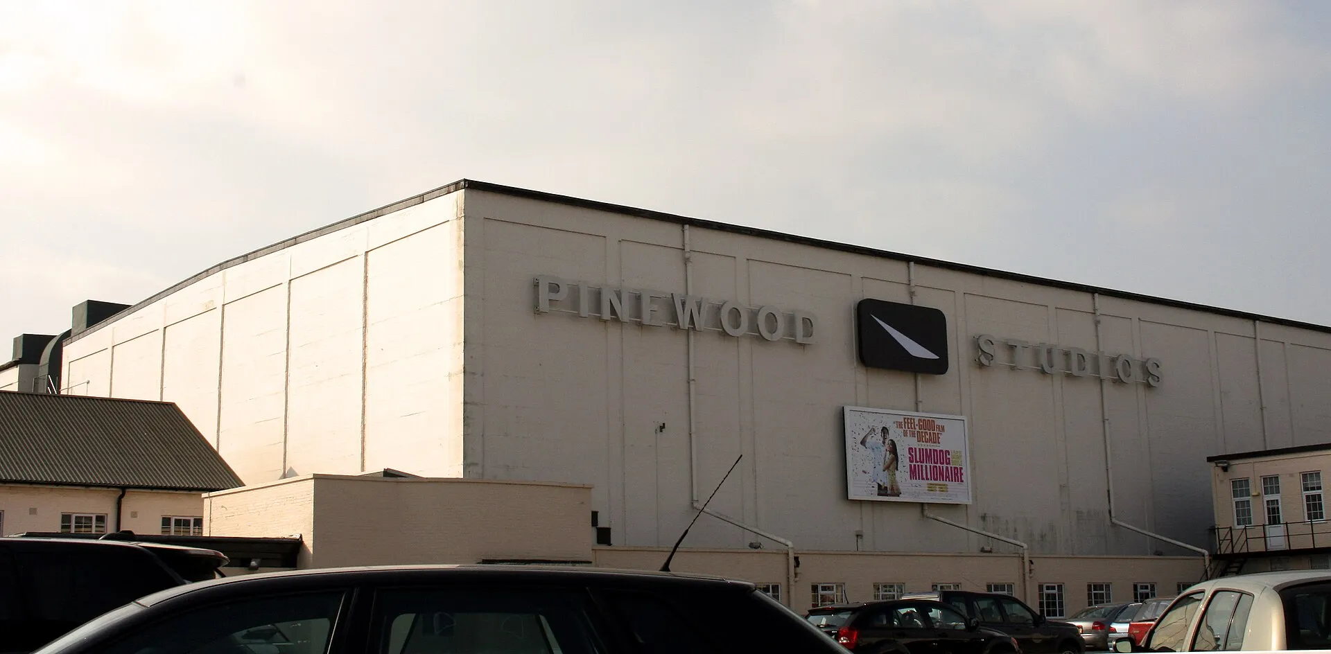 Fassade einer Aufnahmehalle der Pinewood Studios mit dem Schriftzug „Pinewood Studios“ und Parkplatz davor