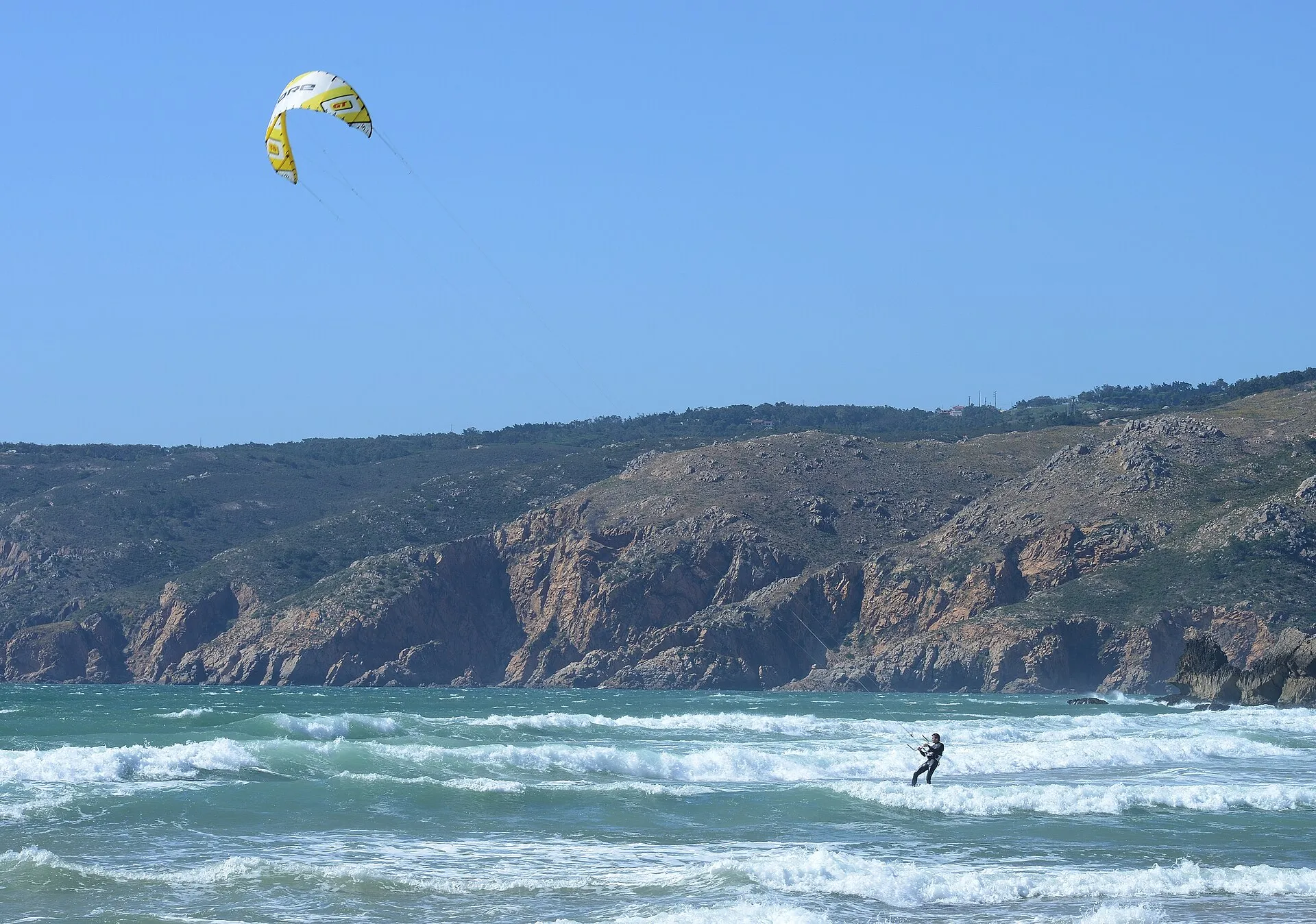 Brandung am Strand von Guincho vor der felsigen Küste der Serra de Sintra, im Wasser ein Kitesurfer