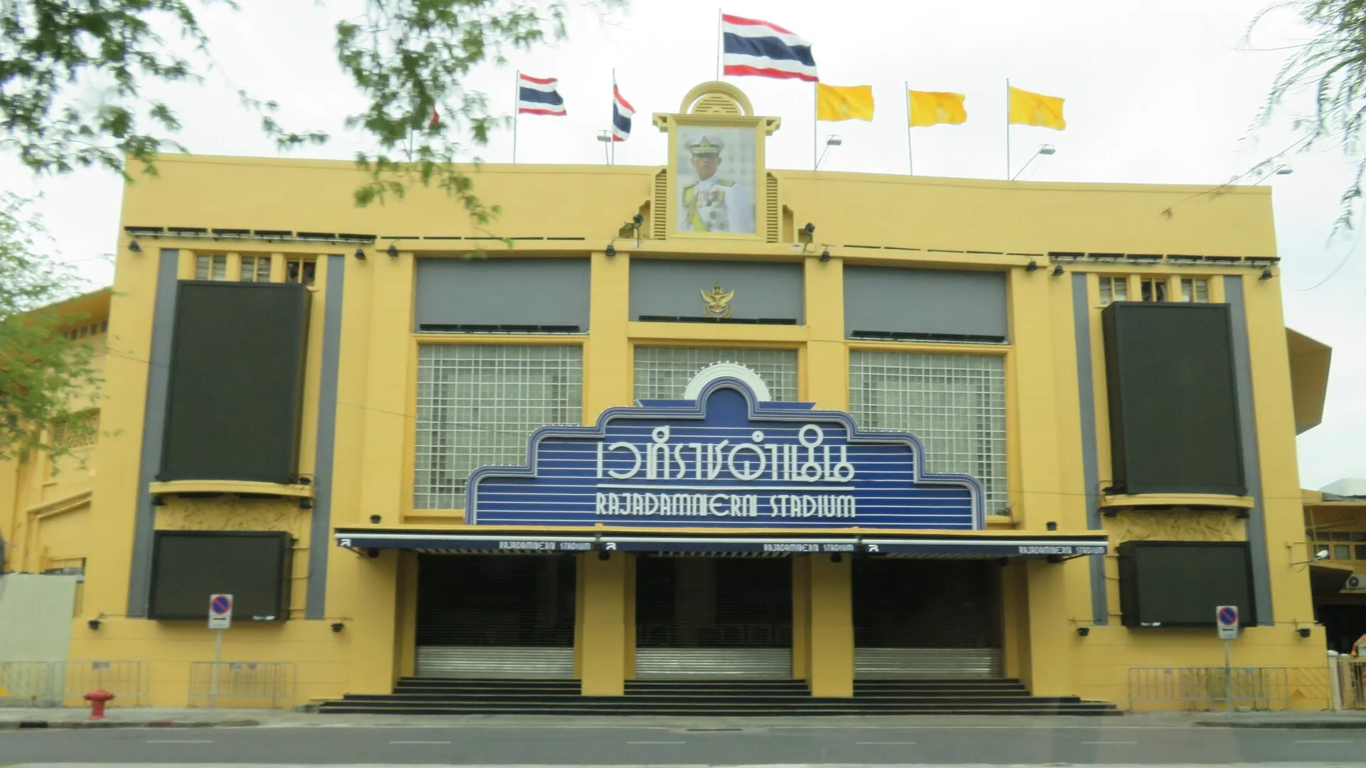 Die gelbe Straßenfassade des Rajadamnern-Stadions in Bangkok mit blauem Namensschild, Königsporträt und thailändischen Flaggen auf dem Dach