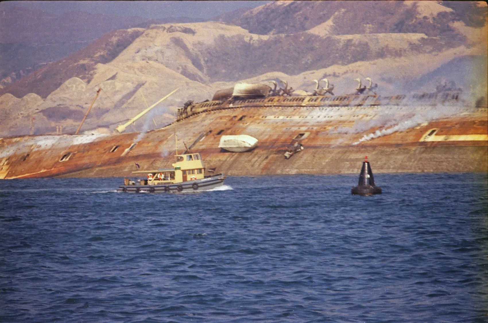 Der ausgebrannte, auf die Seite gekippte und noch rauchende Rumpf des Passagierschiffs Seawise University im Hafen von Hongkong, davor ein Motorboot und eine Boje