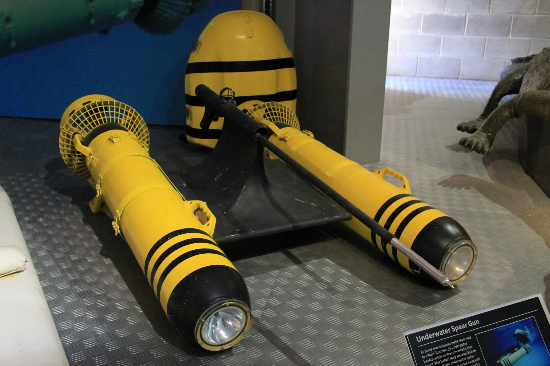 Zwei gelb-schwarz gestreifte Unterwassergeräte mit Scheinwerfer und Gitter vor dem Propeller, dazu ein schwarzer Tauchschlitten und ein gelber Helm in einer Vitrine