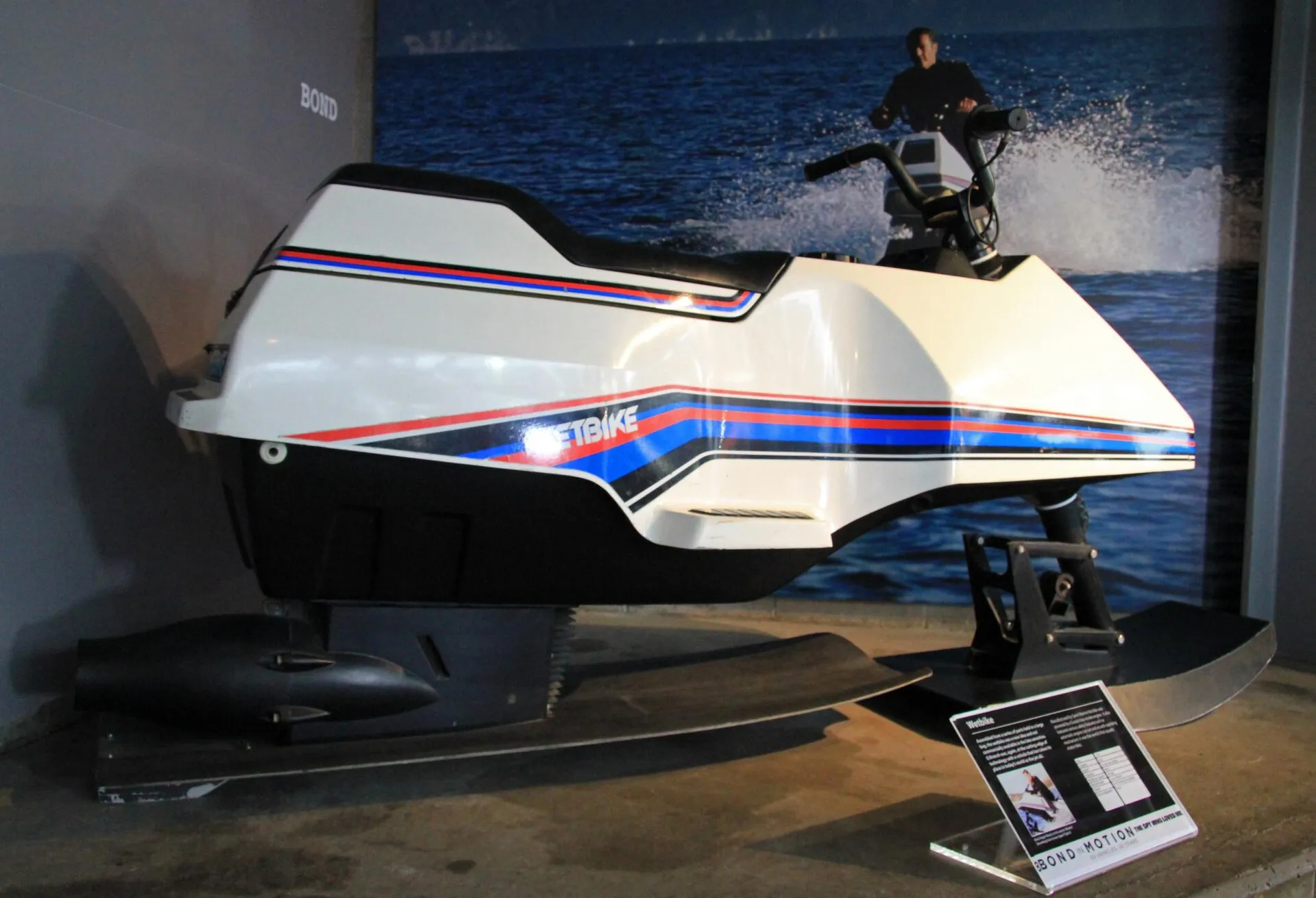Weißes Wetbike mit rot-blauen Zierstreifen auf zwei Kufen im Ausstellungsraum, dahinter ein großformatiges Foto von Bond auf demselben Gerät
